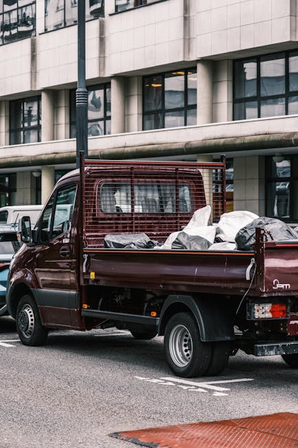 A small burgundy flatbed truck parked on a street in front of a modern office building with large glass windows. The truck's cargo area is loaded with various household items wrapped in plastic, inclu