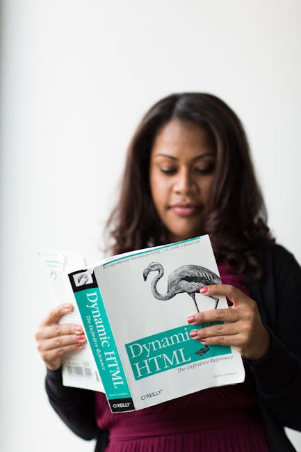 A woman with long dark hair, dressed in a black jacket and burgundy top, is holding a book titled 'Dynamic HTML' with a picture of a flamingo on the cover. She is inside a room with a neutral backgrou